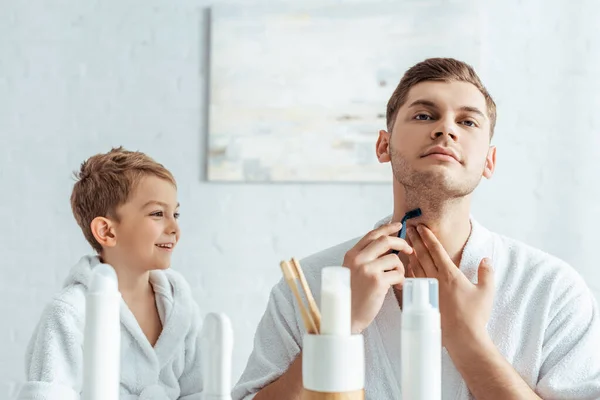 Избирательный фокус молодого человека, бреющего шею рядом с улыбающимся сыном в ванной комнате — стоковое фото