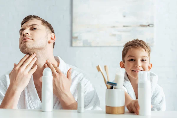 Selective focus of serious man touching face near smiling son in bathroom — Stock Photo