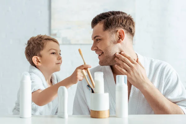 Selective focus of smiling man touching face while adorable son taking toothbrush near toiletries — Stock Photo
