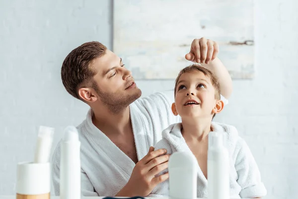 Selective focus of young father touching hair of cheerful son near toiletries in bathroom — Stock Photo