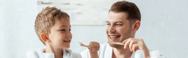 Imagem horizontal de pai e filho felizes olhando um para o outro enquanto escovando os dentes juntos — Fotografia de Stock