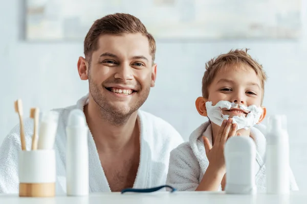 Selective focus of cheerful man looking at camera near adorable son with shaving foam on face — Stock Photo