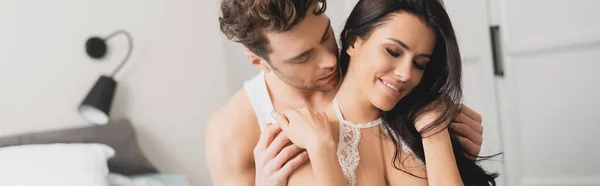 Panoramic crop of handsome man embracing smiling woman in bra in bedroom — Stock Photo