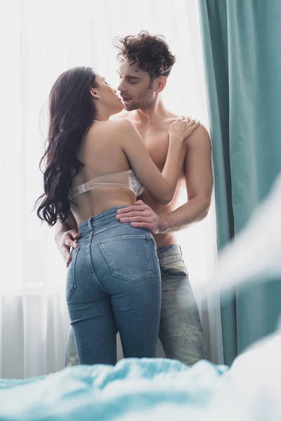 Selective focus of sexy woman kissing muscular boyfriend near window in bedroom — Stock Photo