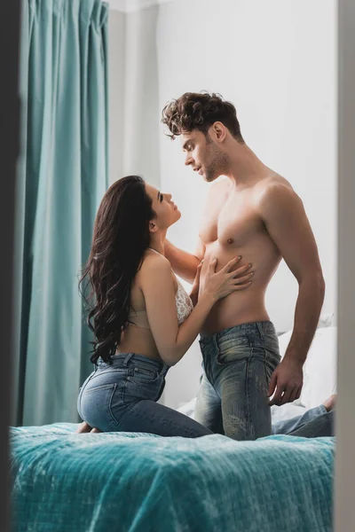 Bel homme musclé regardant femme sensuelle en soutien-gorge et jeans sur le lit — Photo de stock