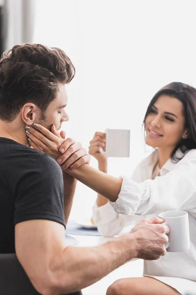 Selective focus of smiling woman touching face of boyfriend while drinking coffee in kitchen — Stock Photo