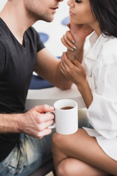 Cropped view of man touching face of sensual girlfriend in shirt while holding cup of coffee — Stock Photo