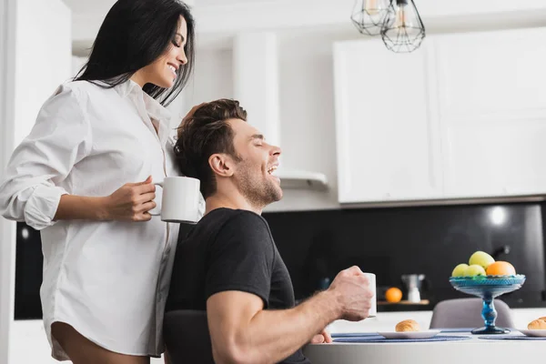 Vue latérale de la femme sexy en chemise tenant tasse de café près copain gai dans la cuisine — Photo de stock