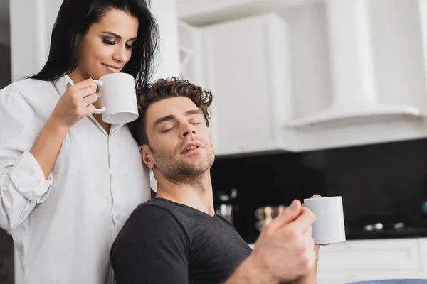 Enfoque selectivo de la hermosa mujer en camisa sosteniendo taza de café cerca de novio guapo en la cocina - foto de stock