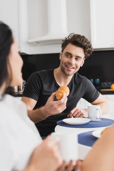 Selektiver Fokus eines gutaussehenden Mannes, der seine Freundin beim Frühstück in der Küche mit einer Tasse Kaffee anlächelt — Stockfoto