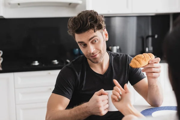 Selective focus of woman flirting with boyfriend holding croissant and cup of coffee in kitchen — Stock Photo