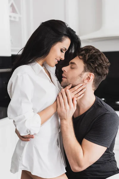 Handsome man hugging smiling girlfriend in shirt in kitchen — Stock Photo