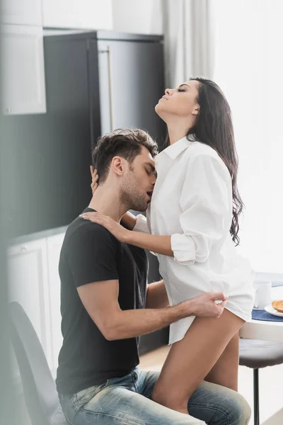 Selective focus of handsome man kissing and undressing sexy girlfriend during breakfast in kitchen — Stock Photo