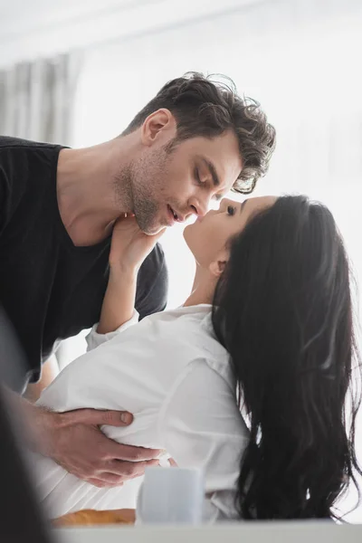 Selektiver Fokus eines hübschen Mannes, der eine schöne Freundin in der Nähe von Kaffee auf dem Küchentisch küsst — Stockfoto