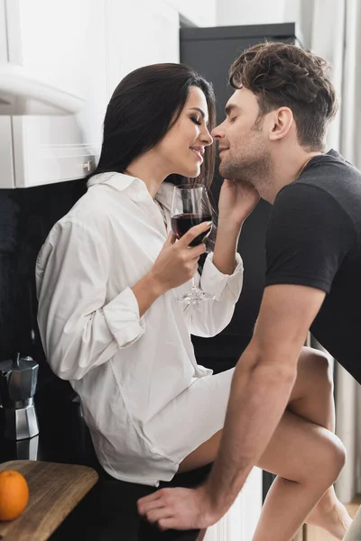 Улыбающаяся женщина в рубашке с бокалом вина и целующимся парнем на кухне — стоковое фото
