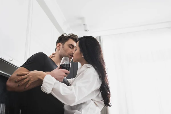 Woman in shirt kissing handsome man with glass of wine in kitchen — Stock Photo