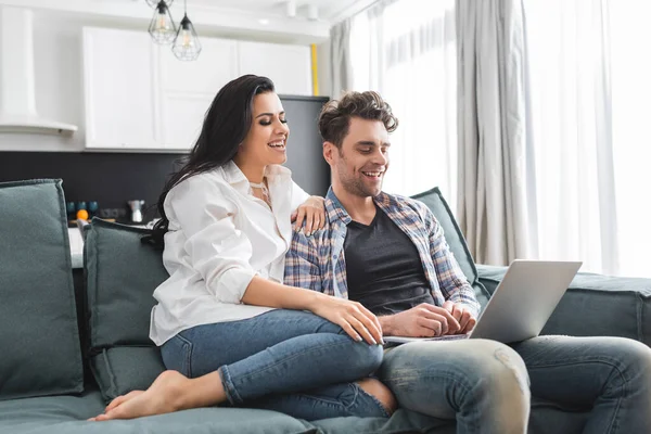 Mujer sonriente sentada cerca de novio guapo usando el ordenador portátil en el sofá en casa - foto de stock