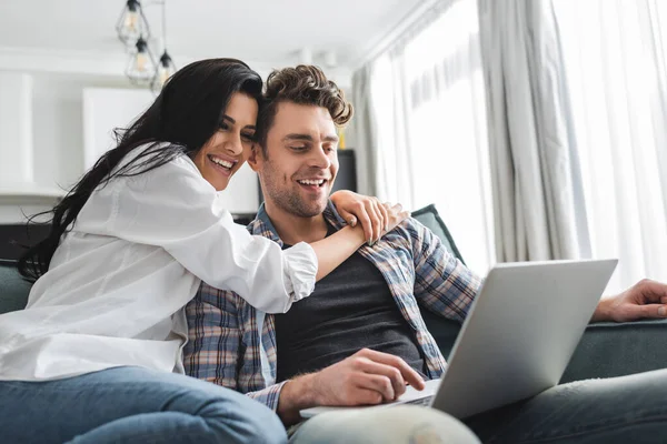 Выборочный фокус улыбающейся женщины, обнимающей парня с помощью ноутбука дома — стоковое фото