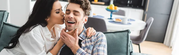 Panoramic crop of beautiful woman kissing smiling boyfriend in living room — Stock Photo