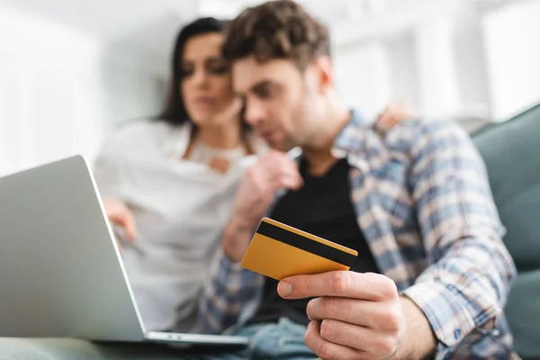 Foco seletivo do homem segurando cartão de crédito ao usar laptop perto da namorada em casa — Fotografia de Stock