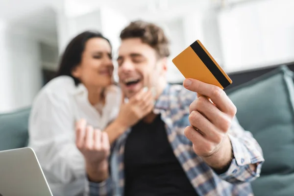 Enfoque selectivo del hombre alegre que sostiene la tarjeta de crédito cerca de la novia y del ordenador portátil en casa - foto de stock