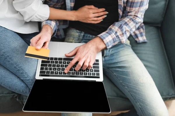 Вид сверху на человека, держащего кредитную карту и использующего ноутбук с экраном-бланком рядом с подругой на диване — стоковое фото