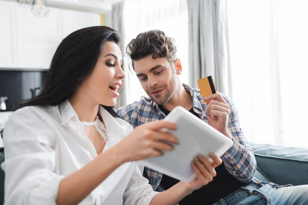 Selective focus of man holding credit card near excited girlfriend showing digital tablet at home — Stock Photo