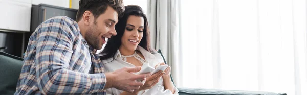 Panoramic crop of smiling couple using smartphones at home — Stock Photo
