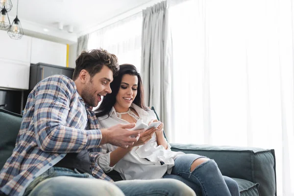 Selective focus of cheerful couple using smartphones on couch in living room — Stock Photo