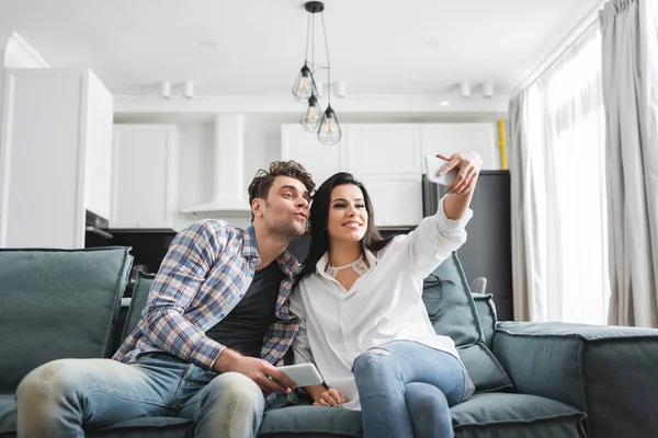 Donna sorridente che si fa selfie con smartphone vicino al fidanzato sul divano di casa — Foto stock