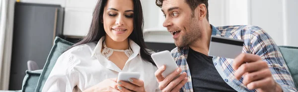 Panoramic shot of positive man holding credit card near girlfriend using smartphone on couch — Stock Photo