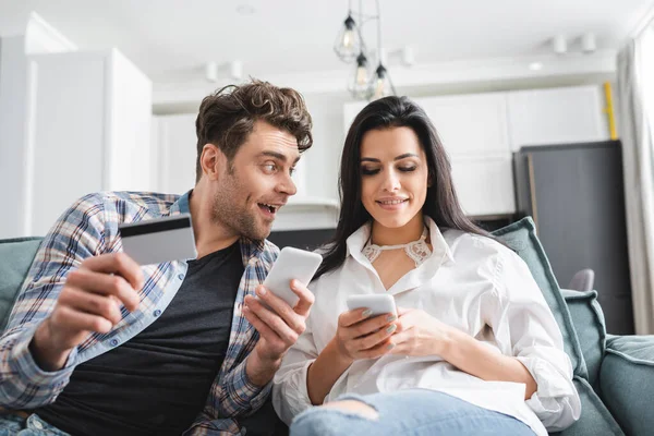 Enfoque selectivo del hombre positivo sosteniendo la tarjeta de crédito y mirando a la novia usando el teléfono inteligente en casa - foto de stock