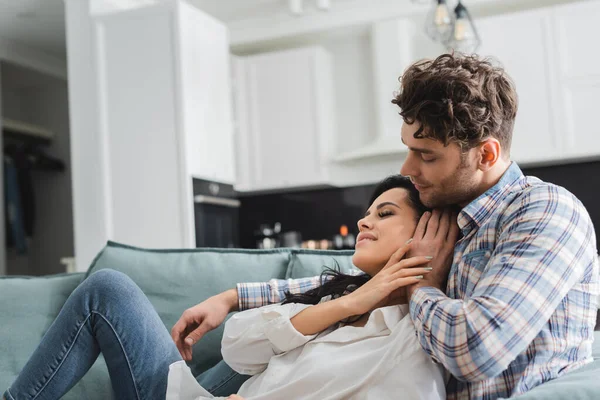 Handsome man hugging and touching hair of beautiful smiling girlfriend on sofa — Stock Photo