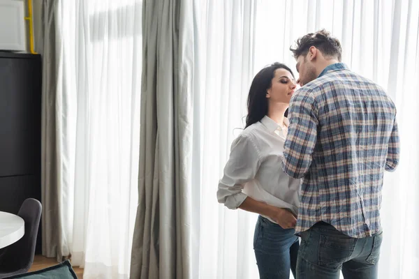 Hombre besando novia sonriente cerca de la ventana en la sala de estar - foto de stock