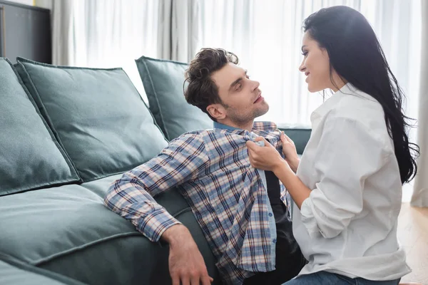 Enfoque selectivo de la mujer sonriente sosteniendo camisa de novio guapo cerca del sofá en la sala de estar - foto de stock