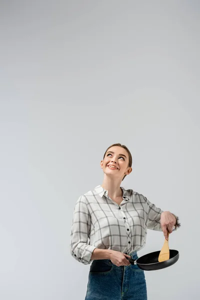 Smiling attractive girl posing like puppet with spatula and frying pan and looking up isolated on grey — Stock Photo