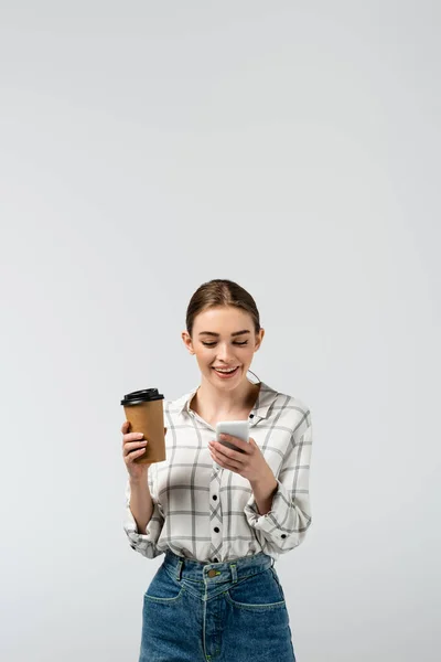 Усміхнена приваблива дівчина використовує смартфон і тримає паперовий стаканчик ізольовано на сірому — стокове фото