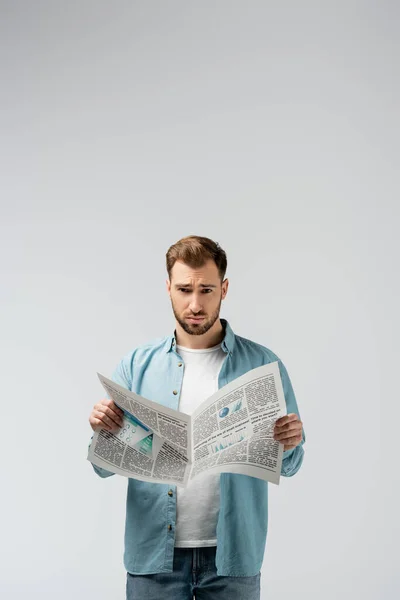 Смущенный молодой человек, читающий газету, изолированную на сером — стоковое фото