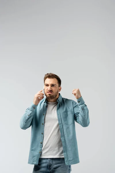 Angry young man talking on smartphone and showing fist isolated on grey — Stock Photo