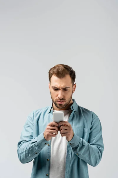 Jeune homme confus utilisant smartphone isolé sur gris — Photo de stock