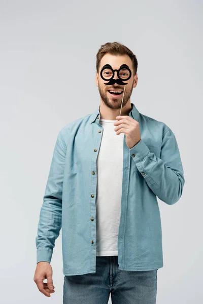 Smiling young man with fake mustache and glasses on stick isolated on grey — Stock Photo