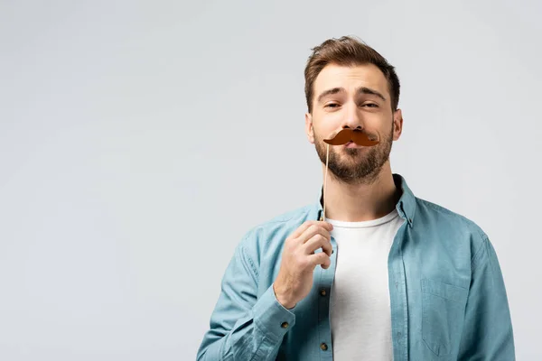 Funny young man with fake mustache on stick isolated on grey — Stock Photo