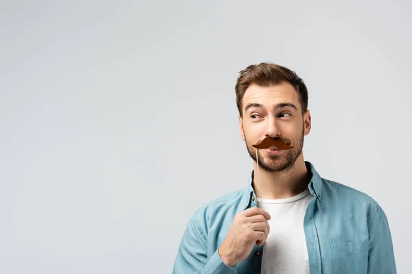 Funny young man with fake mustache on stick looking away isolated on grey — Stock Photo