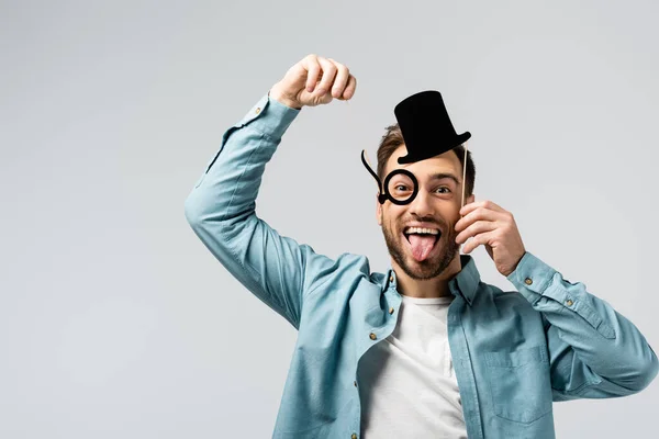 Funny young man with fake hat and Pince-nez showing tongue isolated on grey — Stock Photo