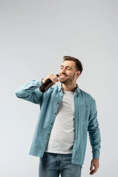 Smiling young man drinking beer from bottle isolated on grey — Stock Photo