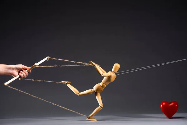 Cropped view of puppeteer holding wooden marionette near red heart on black background — Stock Photo