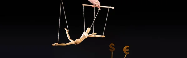 Cropped view of puppeteer holding wooden marionette on strings near currency signs isolated on black, panoramic shot — Stock Photo