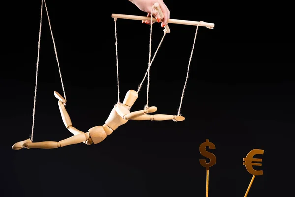 Vue recadrée du marionnettiste tenant une marionnette en bois sur des cordes près de signes monétaires isolés sur du noir — Photo de stock