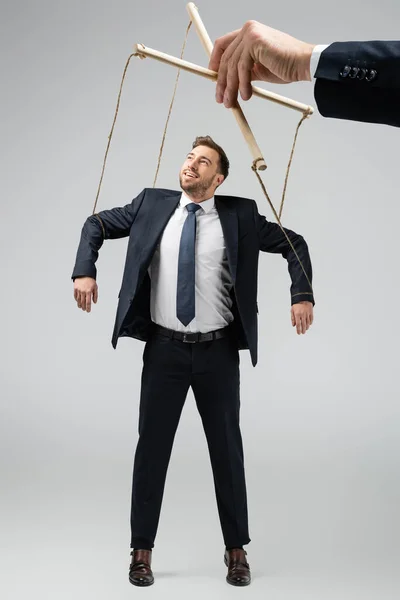 Vista recortada de titiritero sosteniendo sonriente hombre de negocios marioneta en cuerdas aisladas en gris - foto de stock