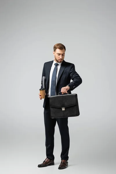 Homem de negócios com mala de couro e copo de papel olhando para relógio de pulso isolado em cinza — Fotografia de Stock